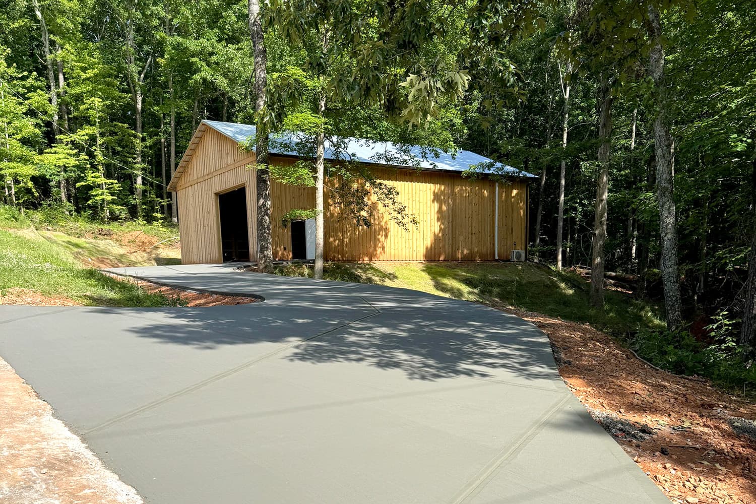 Smooth grey concrete driveway curves toward a vertical-plank wooden barn surrounded by dense green woods.